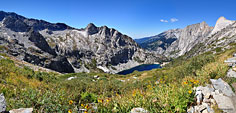 The trail climbs above treeline through grass and shrubs. This 180 degree vista west shows the sheer cliff escarpment known as Valhalla rising above the NW end (right side) of the lake.